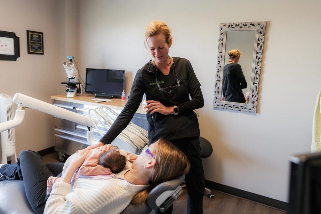In a serene dental clinic situated in Tulsa, Oklahoma, a dedicated biological dentist is seen standing beside a relaxed patient who cradles an infant. The reflection in the wall-mounted mirror captures this health-centered professional, highlighting their unwavering commitment to delivering holistic and comprehensive care while ensuring utmost patient comfort.