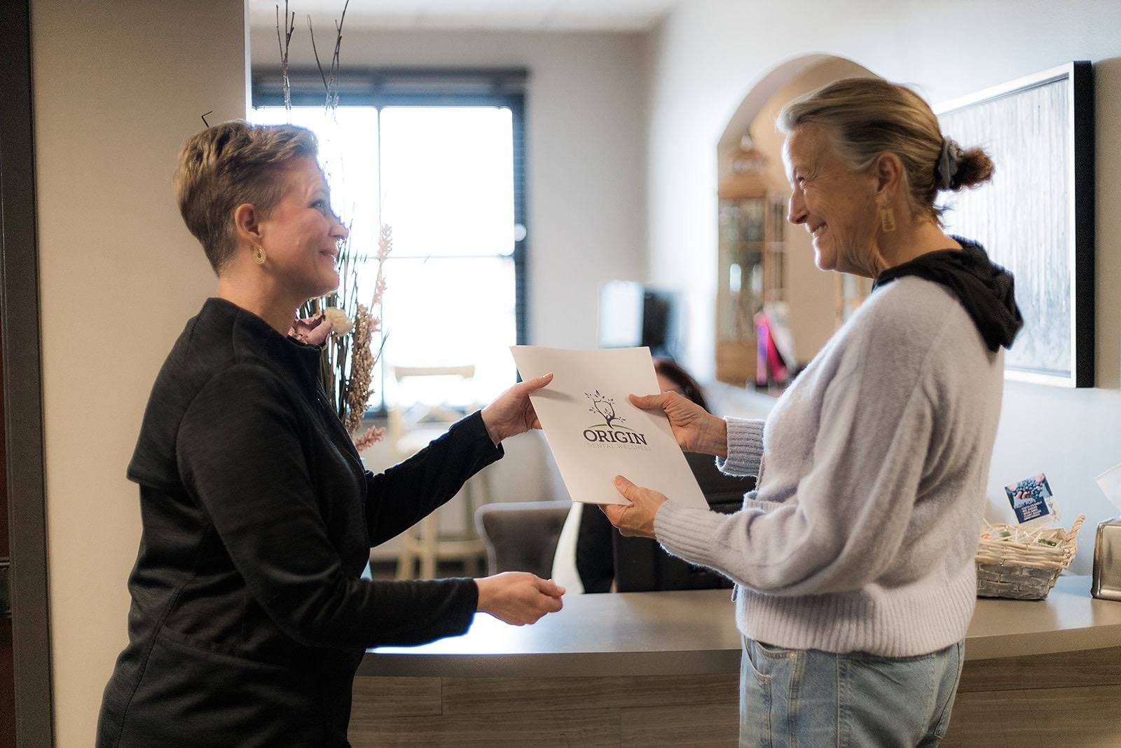 At Origin Dental Wellness in Tulsa, Oklahoma, two women stand by a beautifully adorned counter, exchanging a document titled "Origin." One woman is dressed in a black top, while the other wears a light sweater. Their discussion revolves around the exceptional personalized care provided by Tulsa's leading biological and holistic health-centered dentist. This conversation highlights the unique approach to dental wellness that focuses on integrating overall health with optimal oral care.